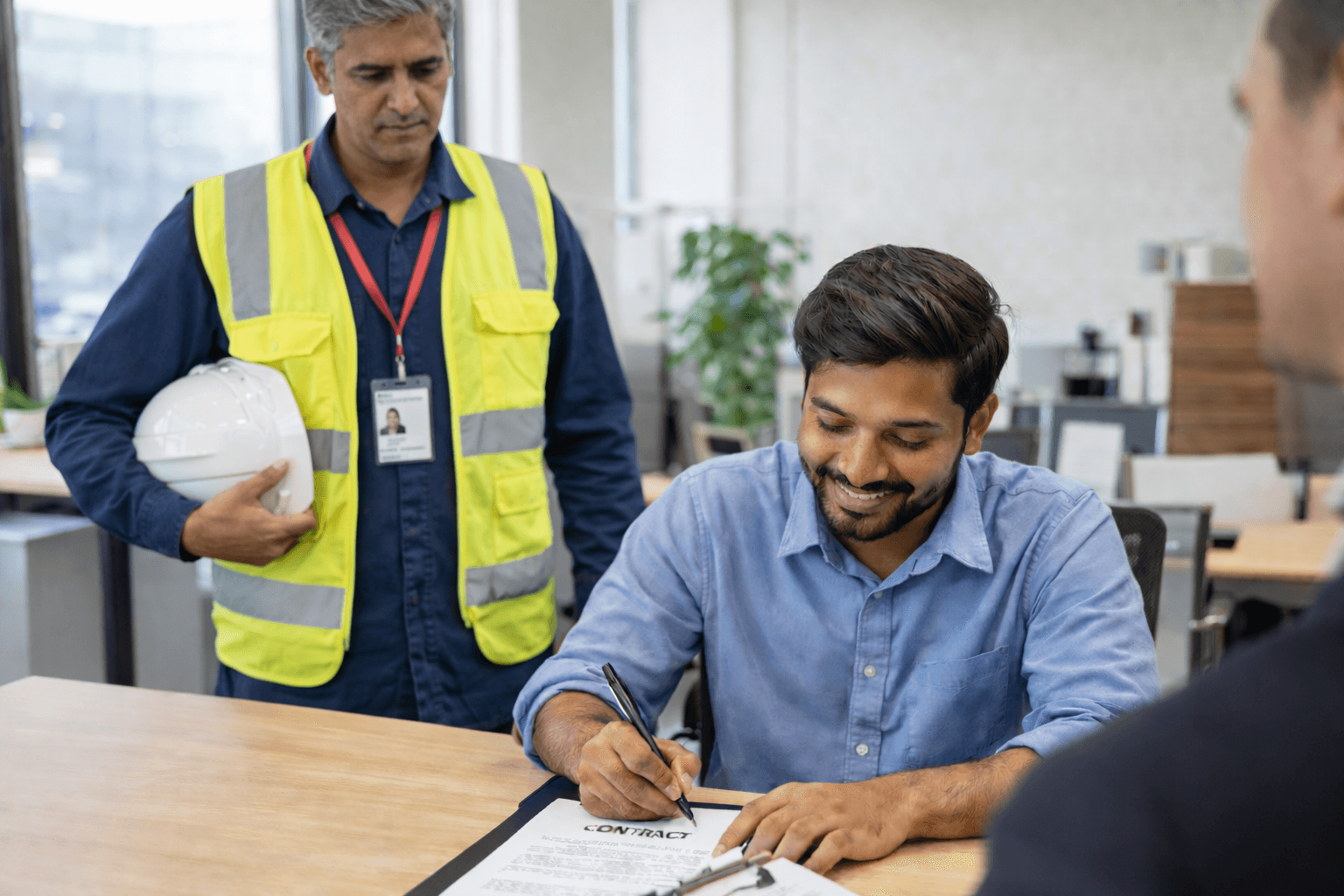 Smiling man signs a contract while a construction worker in a safety vest observes.